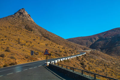 Road leading towards mountains against clear blue sky