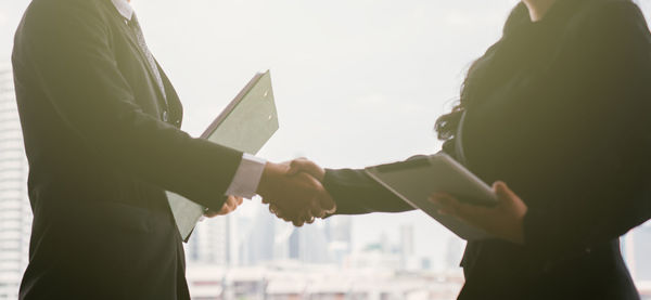 Midsection of business people shaking hands while standing against sky