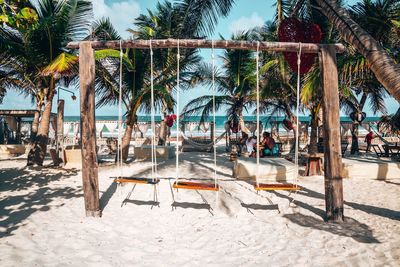 Empty swing and heart shape artwork hanging on tree at beach resort with tourists spending time.