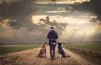 Rear view of man with dogs standing on country road against cloudy sky