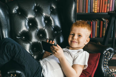 The red-haired boy cuts in an armchair and holds a phone in his hands.