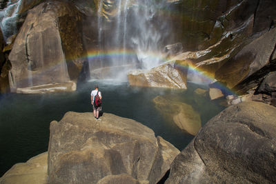 Rear view of man standing on rock formation by sea and rainbow