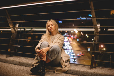 Portrait of young woman sitting on bridge in city at night