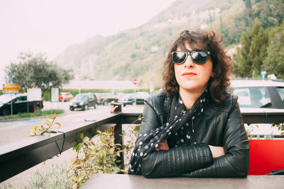 Confident woman wearing sunglasses and jacket while sitting at outdoor cafe