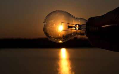 Close-up of hand holding illuminated light bulb against sky during sunset