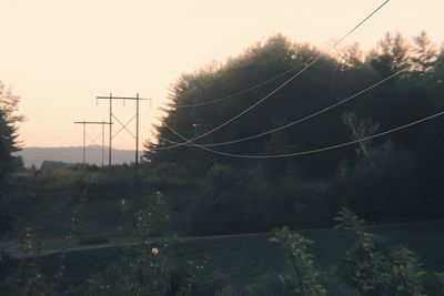 Electricity pylon by trees against sky at sunset