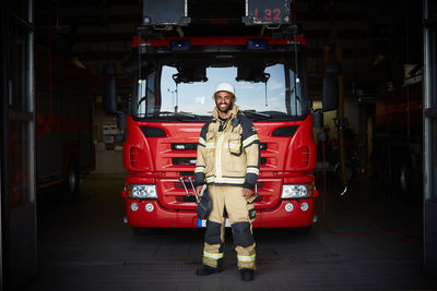Full length portrait of smiling male firefighter standing in front of fire engine at fire station