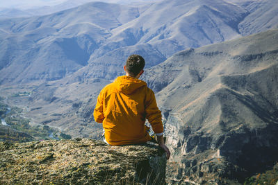 Rear view of man sitting on cliff against mountains