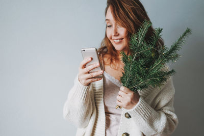 Attractive friendly smiling brunette woman middle age in cozy knitted cardigan using mobile with fir 