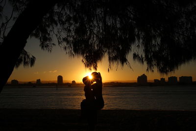 Silhouette couple kissing at riverbank against sky during sunset