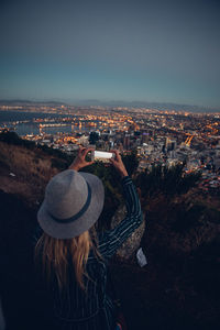 Rear view of woman photographing illuminated cityscape against sky at night