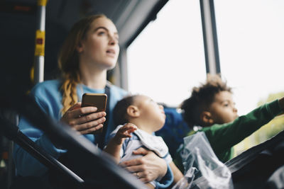 Mother with children holding mobile phone while looking through window from bus