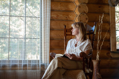 Beautiful blonde woman in beige home clothes sits in a cozy chair and reads a book cozy village life