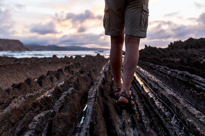 Cropped anonymous male traveler walking on rocky shore near wavy sea under bright cloudy sky at sundown