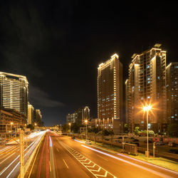 Light trails on road amidst illuminated buildings against sky at night