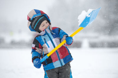 Cute boy holding snow in shovel on field during winter