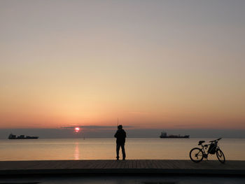 Silhouette man fishing while standing on promenade against sky during sunset