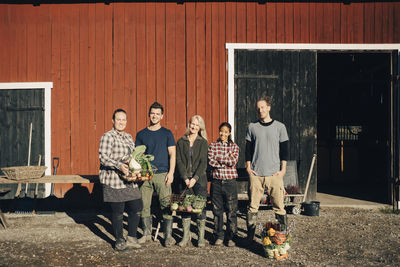 Full length of male and female farmers with organic vegetables standing outside barn