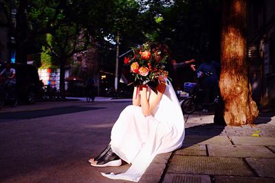 Woman covering her face with flower bouquet sitting on sidewalk