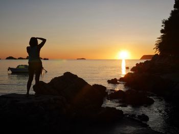 Silhouette woman standing on rock by sea against sky during sunset