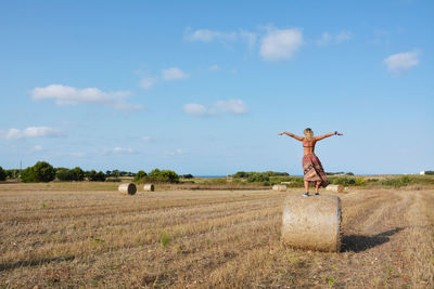 Rear view of woman standing on hay bales at field against sky