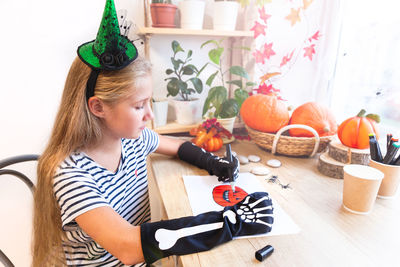 A teenage girl in a green witch hat and black gloves with bones draws a pumpkin by the window.