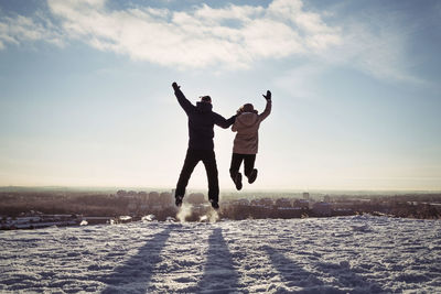Couple holding hands while jumping on snow covered mountain against sky