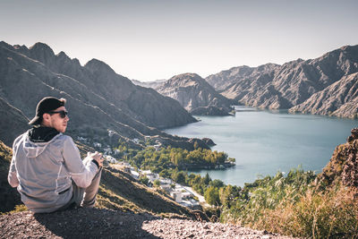 Man sitting on mountain by lake against sky