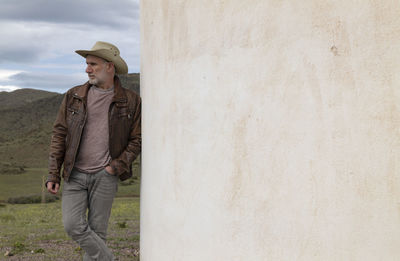 Adult man in cowboy hat standing against white wall in field