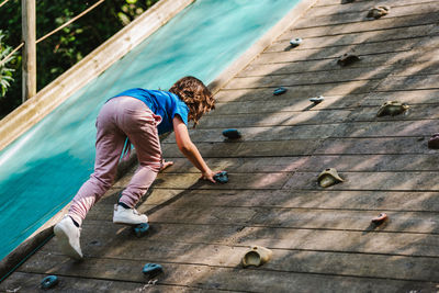 Back view of unrecognizable little girl in casual outfit holding grips while practicing bouldering on climbing wall in lush green park on sunny day