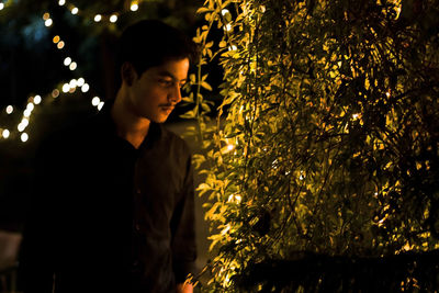 Young man standing by tree with illuminated lights at night