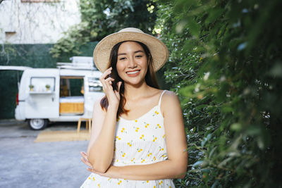 Portrait of smiling young woman wearing hat talking on phone standing by plants