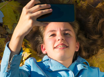 Teenager girl with brunette hair and lying down on the floor covered by yellow leaves while taking a photo with her mobile phone to herself. the girl is smiling to the camera. horizontal phot