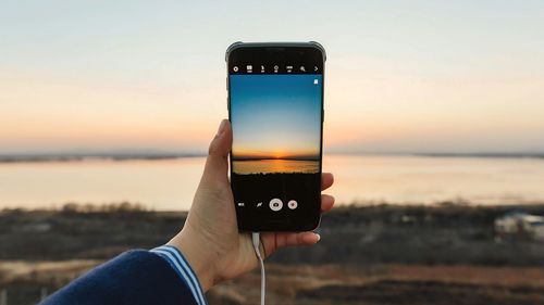 Low section of person holding smart phone against sea during sunset