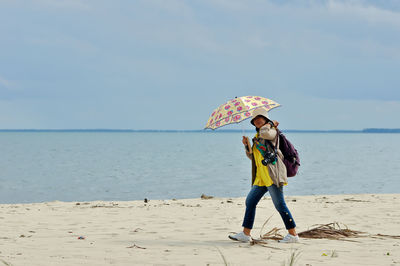 Full length of woman holding umbrella walking on beach against sea