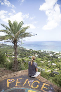 Rear view of woman sitting on rock by sea against sky with palm tree