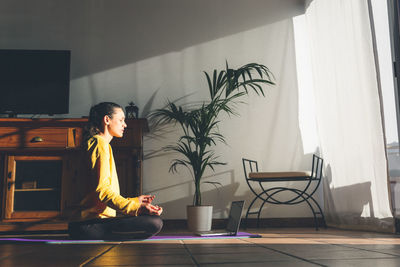 Young caucasian brunette woman practicing yoga at home sitting in lotus pose.