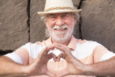 Portrait of senior man wearing hat making heart shape while standing against stone wall
