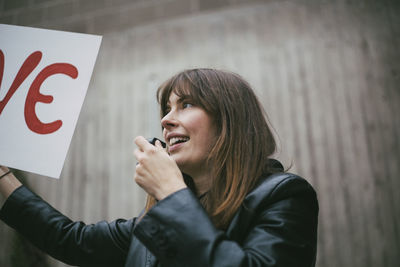 Smiling female activist with whistle holding poster against wall