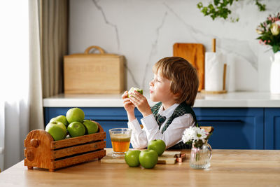 Thoughtful schoolboy is having breakfast with fresh juice and apple waiting for the school bus