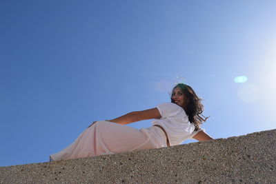 Low angle portrait of smiling woman reclining on retaining wall against clear blue sky