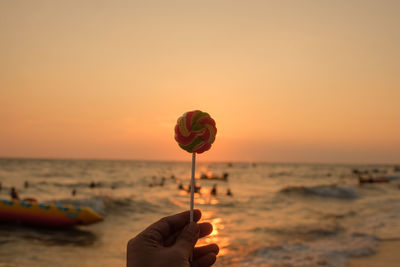 Cropped hand holding lollipop at beach against sky during sunset