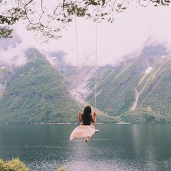 Rear view of woman sitting on swing by lake against mountain