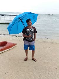 Young man with blue umbrella looking at sky by seashore