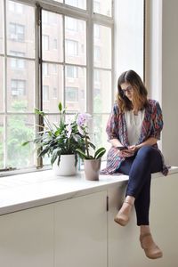 Mid adult woman using mobile phone while sitting on window sill at home