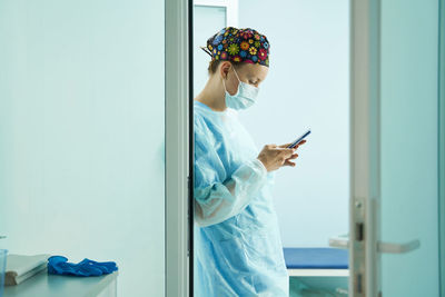 Side view of female doctor in medical uniform and sterile mask text messaging on cellphone in clinic