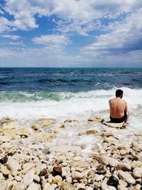 Rear view of shirtless man sitting on rock at beach