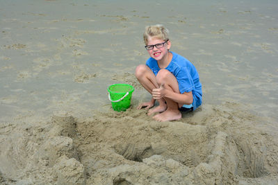 Little blond boy with glasses, playing in the sand on the beach, showing thumbs up