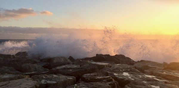 Sea waves splashing on rocks against sky during sunset