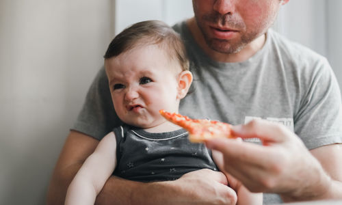 A caucasian baby girl sits in the arms of her dad, who gives her a piece of homemade pizza